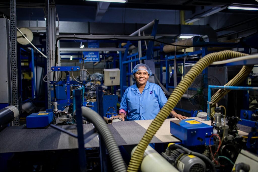 A female worker in a blue uniform smiles while working at a textile factory, surrounded by machinery.